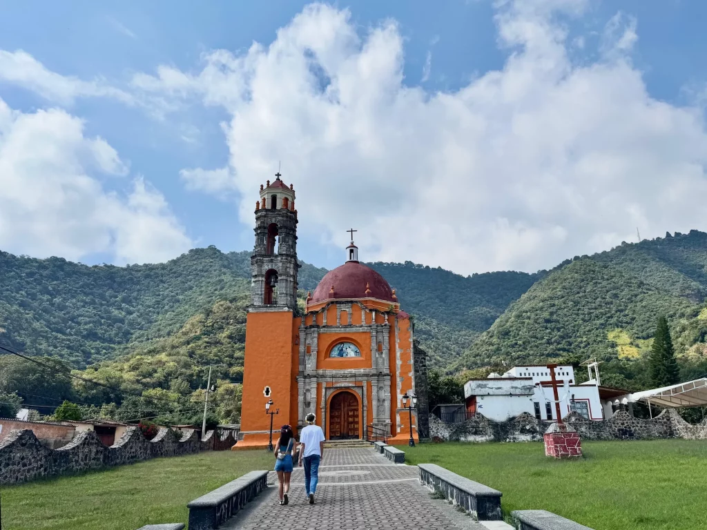 A couple walking towards the front entrance of the Capilla de San Nicolás, the orange church near Malinalco in Mexico on a sunny day