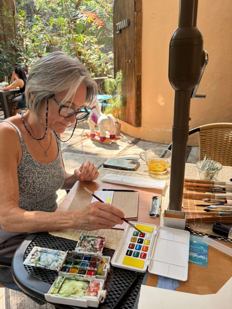 Spanish Immersion student painting in a watercolor painting class outside in a cafe with tropical trees behind